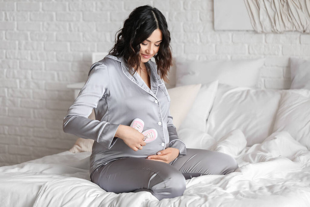 Jonge zwangere vrouw met baby laarzen zitten in de slaapkamer - Foto, afbeelding