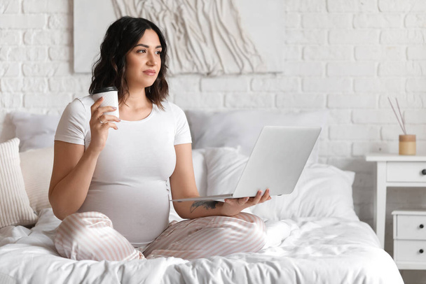Молодая беременная женщина с ноутбуком и чашкой кофе в спальне - Фото, изображение