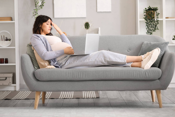 Evdeki kanepede dizüstü bilgisayarla çalışan yorgun ve hamile bir kadın. - Fotoğraf, Görsel