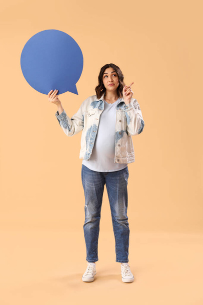 Young pregnant woman with blank speech bubble pointing at something on beige background - Foto, afbeelding