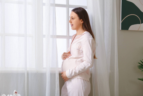 Nuori raskaana oleva nainen pyjamassa lähellä ikkunaa kotona - Valokuva, kuva