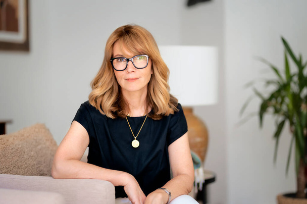 Close-up portrait of attractive middle aged woman relaxing in an armchair at home. Blond haired female wearing eyeglasses and black shirt. - Foto, afbeelding