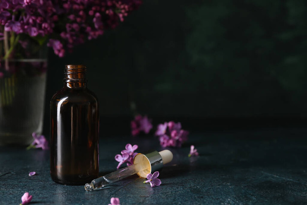 Bottle of cosmetic oil with beautiful lilac flowers on dark table - Фото, изображение