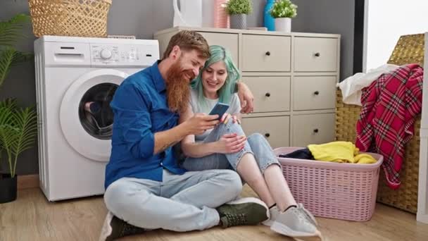Man and woman couple using smartphone sitting on floor at laundry room - Footage, Video