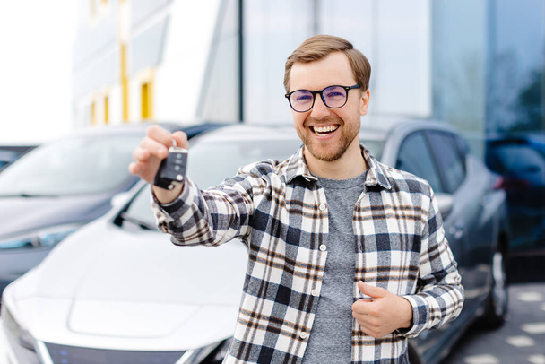 Emocionální chlap drží klíč v ruce a usmívá se do kamery. Muž ukazující klíč od svého nového elektrického auta. Mladý muž kupující auto v prodejně. - Fotografie, Obrázek