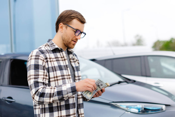 Young man holding money in hand standing front car and prepare pay by installments - insurance, loan and buying car concept insurance. Buy and down payment a car. - Photo, Image