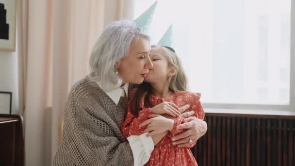 Grootmoeder en kleindochter knuffelen op het verjaardagsfeest kijkend naar de camera. Oudere vrouw en meisje kletsen en zoenen op het feest. Hoge kwaliteit 4k beeldmateriaal - Video