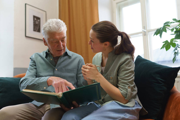 Grandfather with his granddaughter looking at family photo album. Nostalgic senior man showing photos of himself from his youth, remebering old days through pictures. - 写真・画像