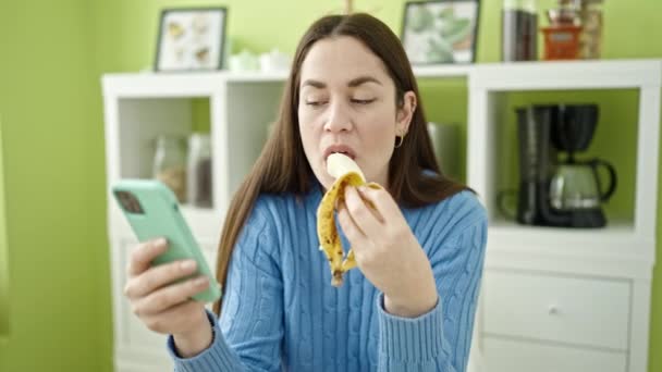 Mladý kavkazský žena jíst banán pomocí smartphone v jídelně - Záběry, video
