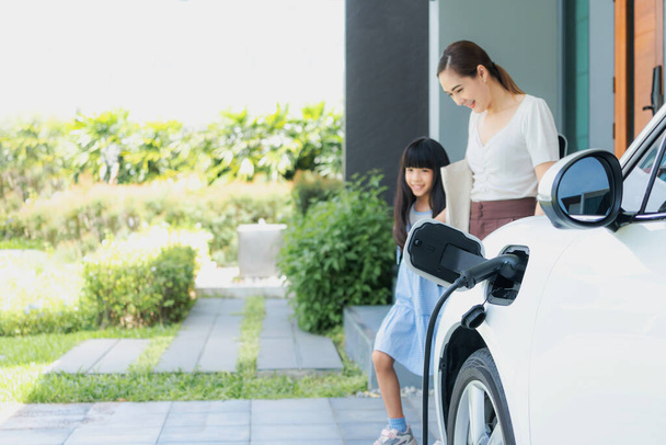 Focus electric vehicle recharging at home charging station plugged in with EV charger device with blurred background of progressive mother and daughter walking as concept for sustainability of energy. - Photo, image