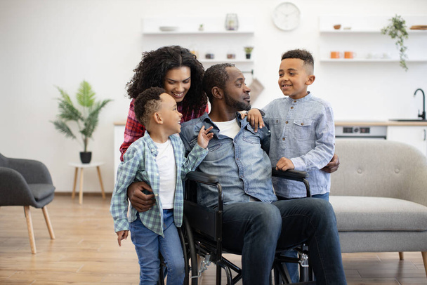 Delighted multicultural adults and playful boys staying in spacious living room of apartment during weekend. Joyful sons talking about sporting success at school to smiling mom and dad in wheelchair. - Foto, Imagen