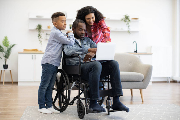 Cheerful multiracial man using wheelchair holding laptop on knees while pretty woman and funny boy standing behind. Loving family of three choosing movie for watching together on Sunday at home. - Valokuva, kuva