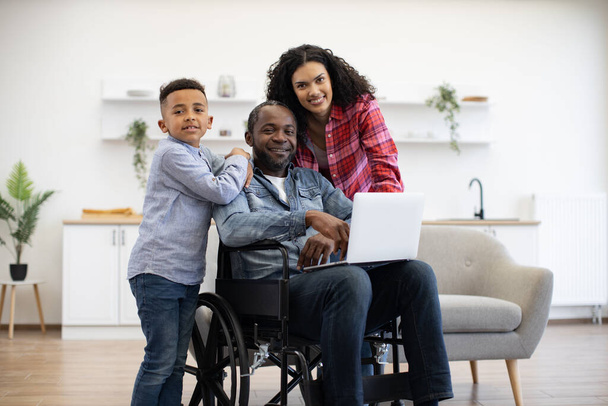 Cheerful multiracial man using wheelchair holding laptop on knees while pretty woman and funny boy standing behind. Loving family of three choosing movie for watching together on Sunday at home. - Zdjęcie, obraz