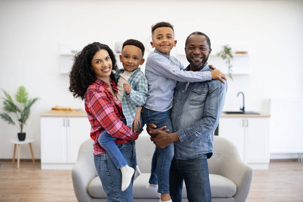 Portrait of active african american spouses holding well-behaved children in arms while feeling comfortable indoors. Loving family man and wife planning enjoyable activities with sons in apartment. - Φωτογραφία, εικόνα