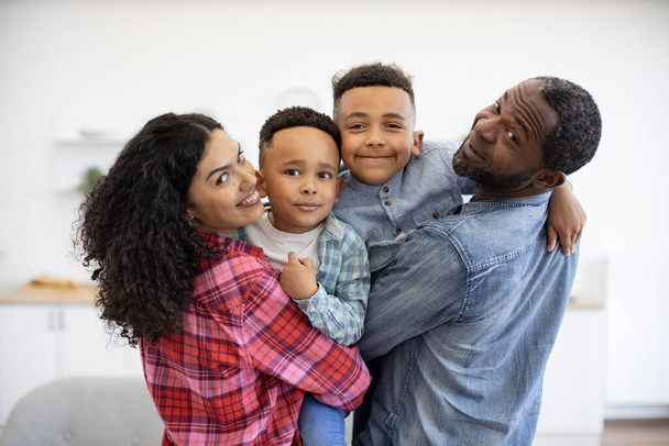 Portrait of smiling multiethnic man and woman with cute kids in arms looking over shoulder in apartment interior. Delighted parents and cute sons displaying affection for each member of loving family. - 写真・画像