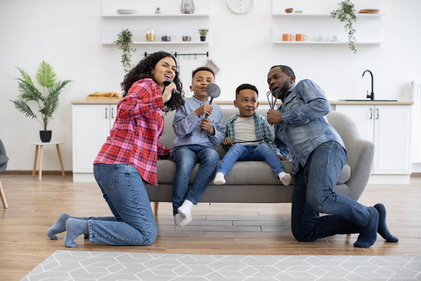 Joyful multicultural woman and man standing on knees near sofa with boys on it while pretending to take part in karaoke. Artistic parents and playful sons singing with kitchen utensils as microphones. - Foto, afbeelding