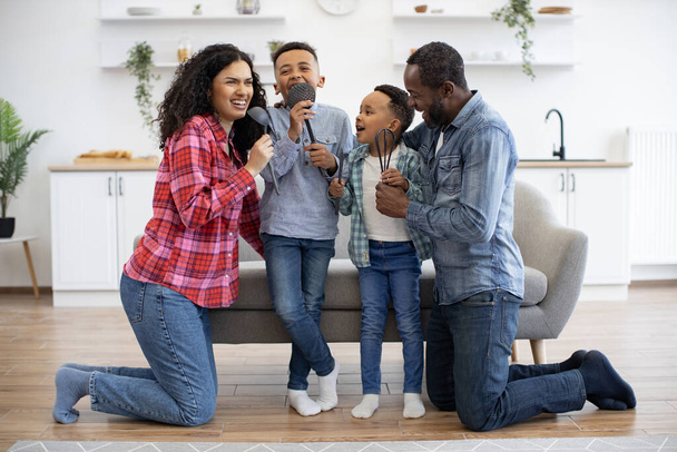 Joyful multicultural woman and man standing on knees near sofa with boys on it while pretending to take part in karaoke. Artistic parents and playful sons singing with kitchen utensils as microphones. - Foto, Imagen