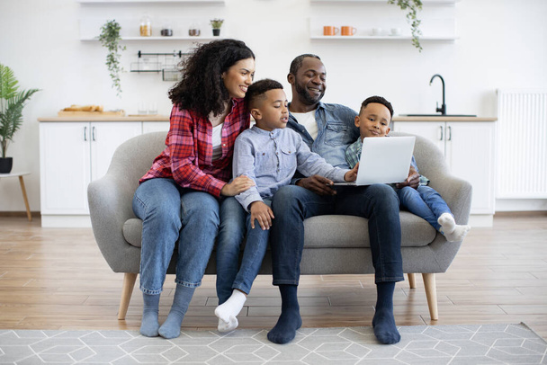 Full length view of multiracial dad holding laptop while joyful four-person family sitting on soft sofa at home. Interested children watching movie with smiling parents in lounge of spacious flat. - Fotografie, Obrázek