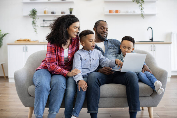 Affectionate african american mom tickling one of cute sons while happy father showing video to another boy. Cheerful parents and children using wireless laptop while having fun at home. - Fotografie, Obrázek