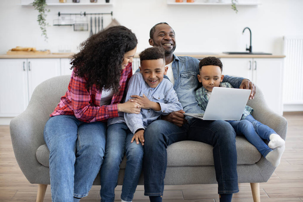 Affectionate african american mom tickling one of cute sons while happy father showing video to another boy. Cheerful parents and children using wireless laptop while having fun at home. - Fotografie, Obrázek