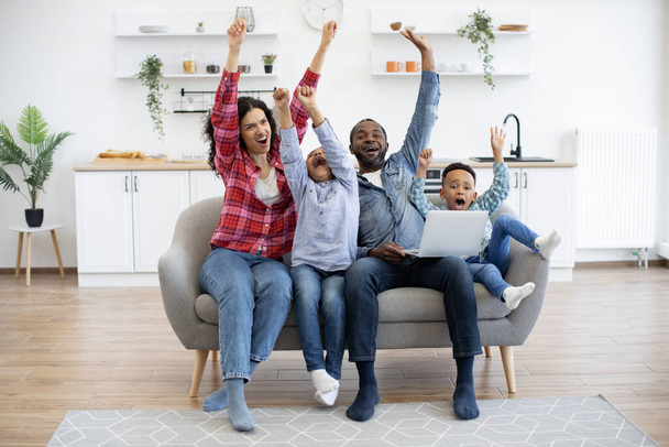Emotional multiracial people raising arms high in air while watching online football match on laptop at home. Excited spouses and little sons expressing real fans joy after winning goal at finals. - Foto, imagen