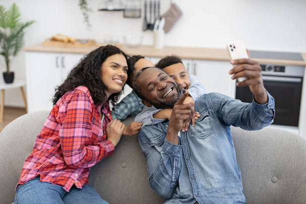 Positive multicultural family of four using modern smartphone while resting in living room. Adorable sons embracing parents while dad taking selfie via mobile webcam in room interior. - Foto, afbeelding