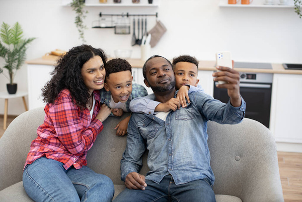 Positive multicultural family of four using modern smartphone while resting in living room. Adorable sons embracing parents while dad taking selfie via mobile webcam in room interior. - Photo, Image