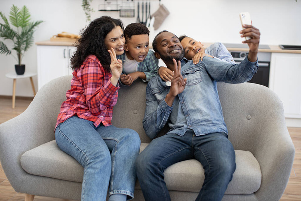 Smiling multicultural father with phone and mother showing V sign while energetic kids standing behind sofa in lounge of home. Happy couple with sons getting self-portrait via mobile app at leisure. - Photo, image