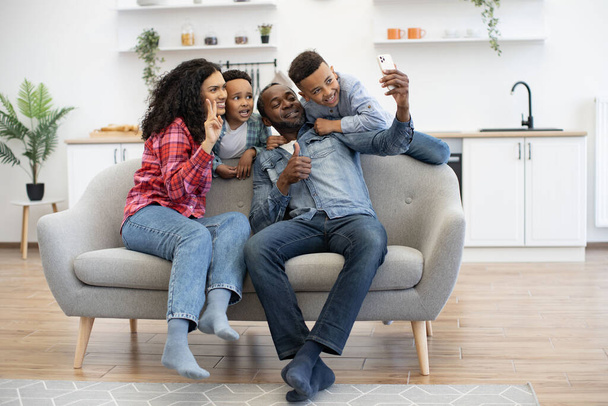 Полный обзор африканских американских родителей, дающих большой палец вверх и знак V во время фотографирования на смартфоне с двумя мальчиками. Веселые взрослые и дети спасают счастливые моменты времяпрепровождения вместе. - Фото, изображение