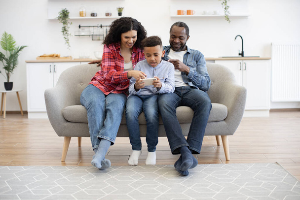 Pretty multiethnic woman sitting next to attractive man and cute boy with smartphone in hands in modern living room. Cheerful mother scrolling photos from last birthday party on sons gadget. - 写真・画像