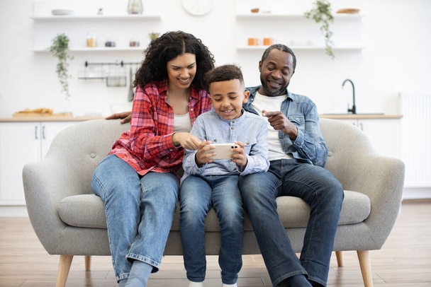 Inquisitive multiracial couple pointing fingers on phone screen held by calm little boy while relaxing on couch in lounge. Helpful parents giving advice about online video game after tasty dinner. - Fotó, kép