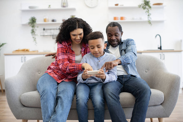 Inquisitive multiracial couple pointing fingers on phone screen held by calm little boy while relaxing on couch in lounge. Helpful parents giving advice about online video game after tasty dinner. - Foto, Imagen