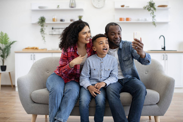Positive african american spouses and active son having online call over cell phone held by father at home. Surprised people being involved in pleasant conversation with relatives via modern gadget. - Foto, Imagem