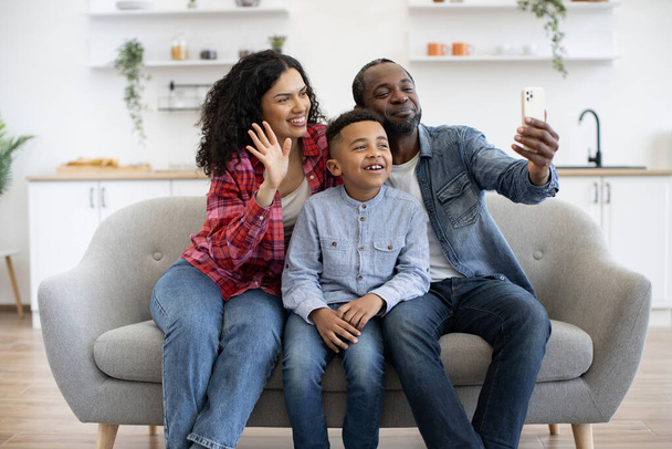 Charming multiracial woman waving hello while proud man with smiling boy staring at webcam of smartphone indoors. Affectionate family of three greeting grandma using video call application on weekend. - Photo, Image
