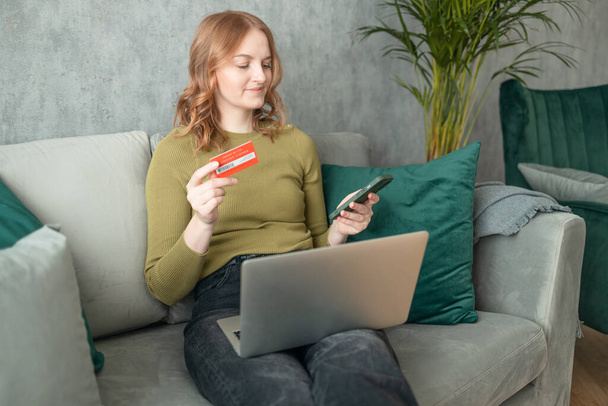 Young woman holding credit card and using laptop computer sitting on couch at home. Pretty relaxed lady holding smartphone buying in ecommerce shop, watching videos online, texting or checking social - Photo, Image