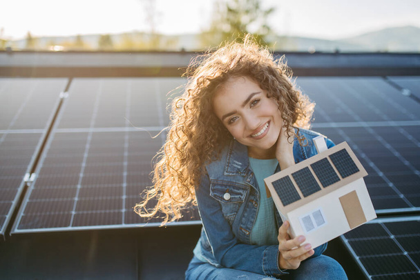 Portrait of young woman, owner on roof with solar panels,holding model of house with photovoltaics. - Valokuva, kuva