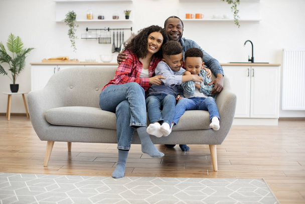 Full length portrait of multiethnic kids with female resting on couch while happy man embracing everyone in kitchen. Playful brothers and merry spouses being involved in relaxing pastime at home. - Foto, Imagen