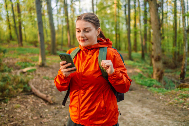 Side view of young female traveler with backpack and in warm wearing standing in the spring forest looking at phone screen. Women on hiking trail. Wooden path in forest. National Park in Poland - Фото, изображение