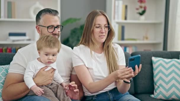 Familia de madre, padre y bebé tomando selfie sentado en el sofá en casa - Imágenes, Vídeo