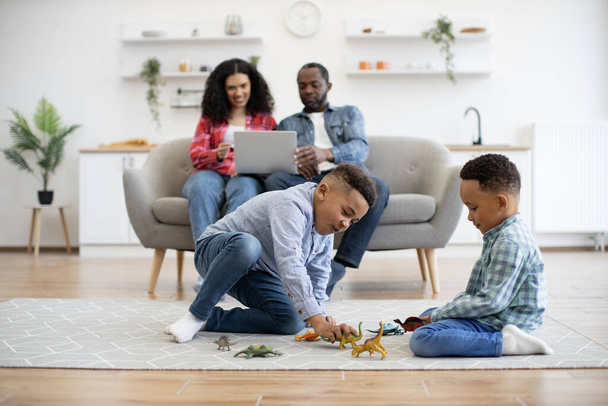 Focus on two multiracial brothers sitting on carpet in living room while parents using laptop on sofa in home interior. Cheerful kids playing with toy dinosaurs while adults watching online TV show. - Photo, image