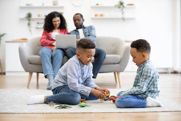 Focus on two multiracial brothers sitting on carpet in living room while parents using laptop on sofa in home interior. Cheerful kids playing with toy dinosaurs while adults watching online TV show. - Photo, Image