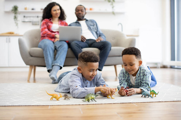 Selective focus of playful multicultural kids lying on floor coverd with carpet while relaxed spouses sitting on sofa with laptop. Inquisitive boys being engaged in adventurous game with dinosaurs. - Photo, Image