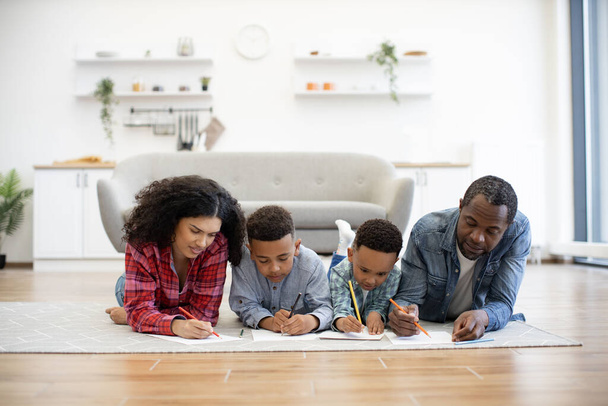 Four african american people using colored pencils while lying on warm wooden floor in kitchen interior. Focused mother and father helping little sons with drawing during summer vocations at home. - Foto, Bild