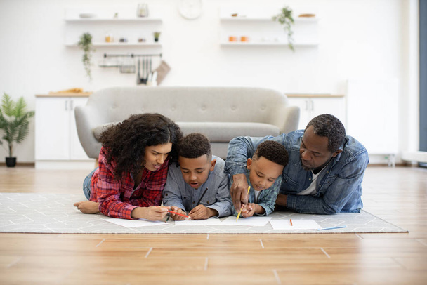 Four african american people using colored pencils while lying on warm wooden floor in kitchen interior. Focused mother and father helping little sons with drawing during summer vocations at home. - Фото, изображение