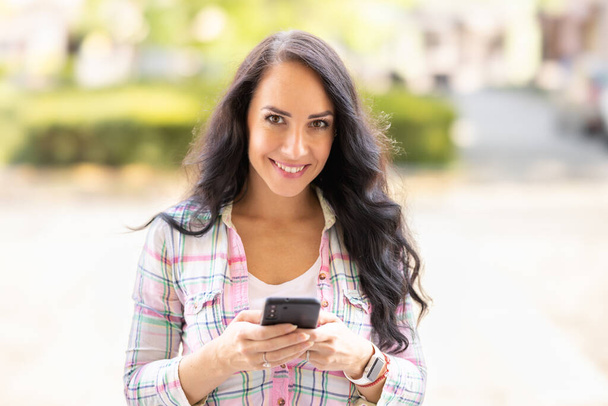 A smiling brunette is writing messages on her mobile phone. A young woman reads received messages and writes back on a smartphone. - Photo, image