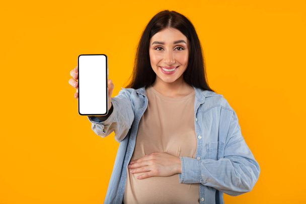 Happy european pregnant lady with big belly showing smartphone with white blank screen on yellow studio background, mockup. Expecting baby, communication app, website recommendation, blog - Photo, Image