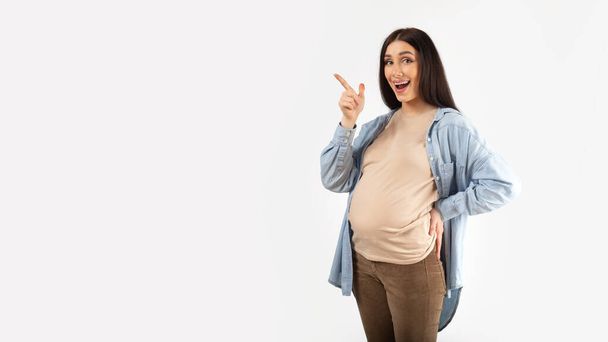 Реклама о беременности. Возбужденная беременная женщина, указывающая в сторону на пространство для копирования на белом фоне, панорама. Красивая отхаркивающая дама, показывающая место для вашего дизайна или рекламы - Фото, изображение