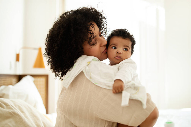 Happy sweet black curly millennial mother with towel kissing little baby, enjoy motherhood in bedroom. Child care, parenthood and family at home, safe and comfort for newborn - Foto, imagen