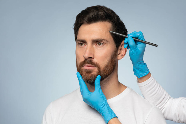 Age-defying treatments. Plastic surgeon drawing contours with black pen on male patient face before surgery, middle aged bearded man getting beauty treatment - Photo, Image