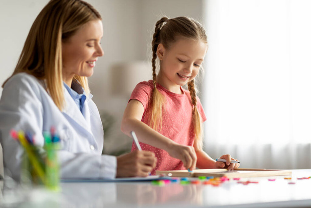 Art Therapy For Kids. Cute Little Girl Painting On Draw Board During Meeting With Child Development Specialist, Therapist Lady Observing Kid Patient Making Learning Activities, Closeup - 写真・画像